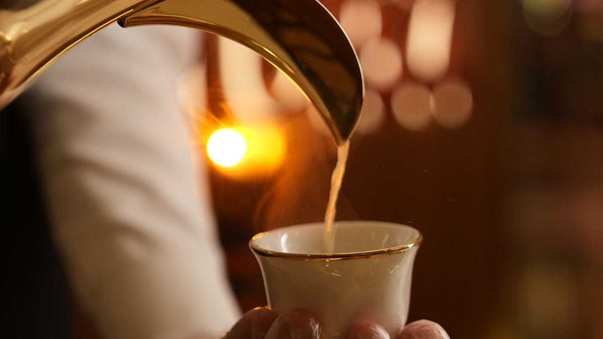 A close-up of a traditional gold Arabic coffee pot pouring hot coffee into a small white cup held by a person wearing a white uniform and gloves, with a warm, blurred background.